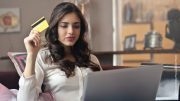 Junge Frau mit dunklen Haaren und einem silbernen Laptop hält eine Kreditkarte hoch